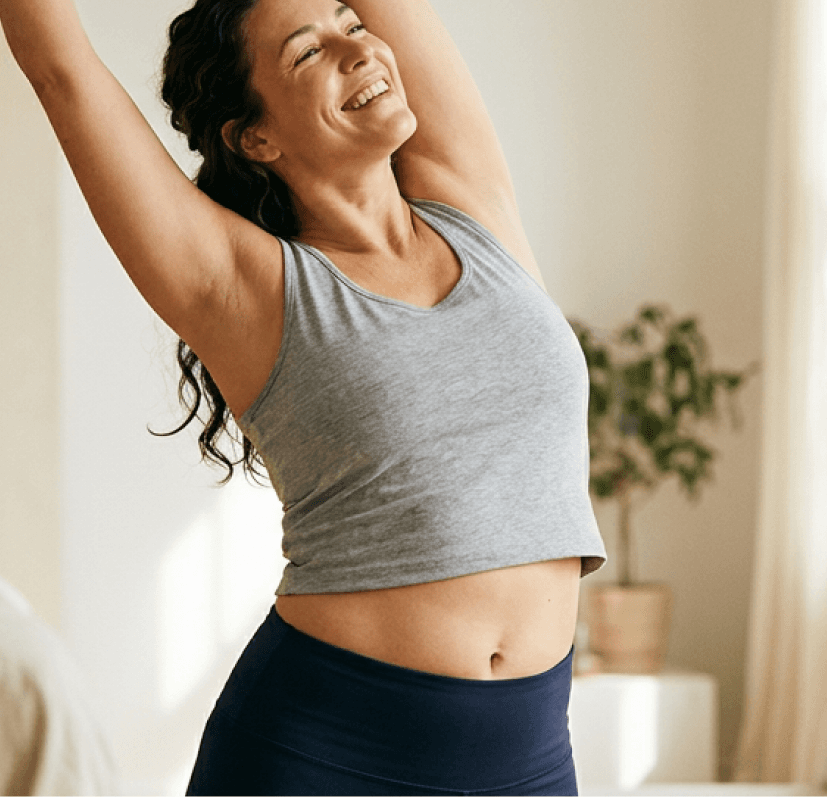 A smiling woman in activewear stretching her arms overhead in a bright room