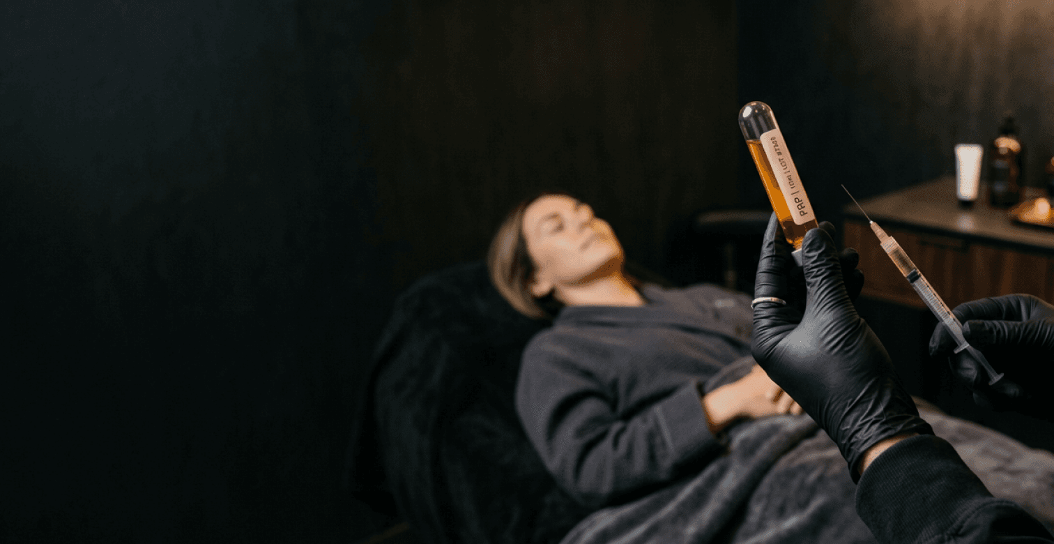 A clinician in black gloves holding a vial of platelet-rich plasma and a syringe beside a resting patient