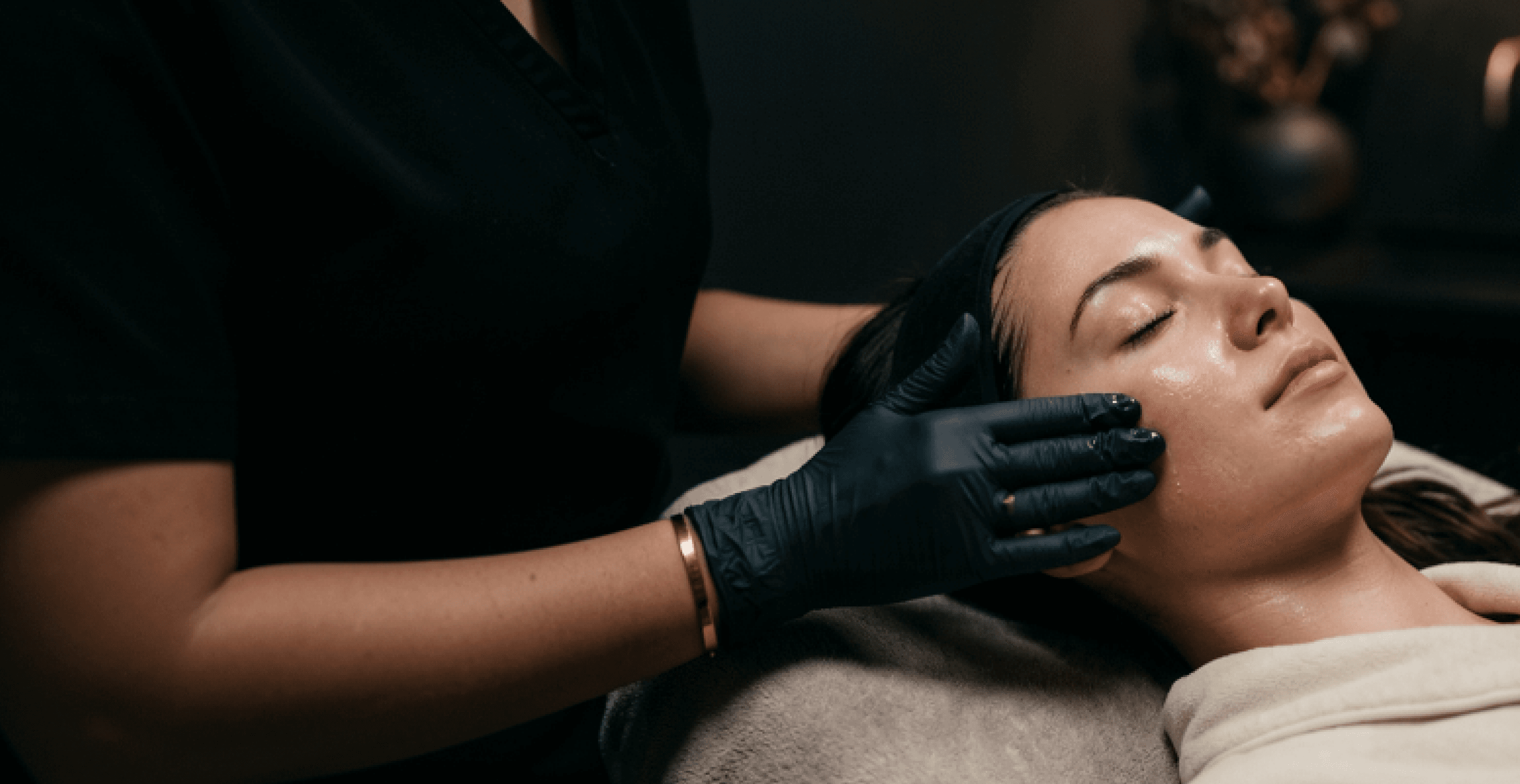 A patient receiving a facial treatment from a gloved clinician in a dark spa room