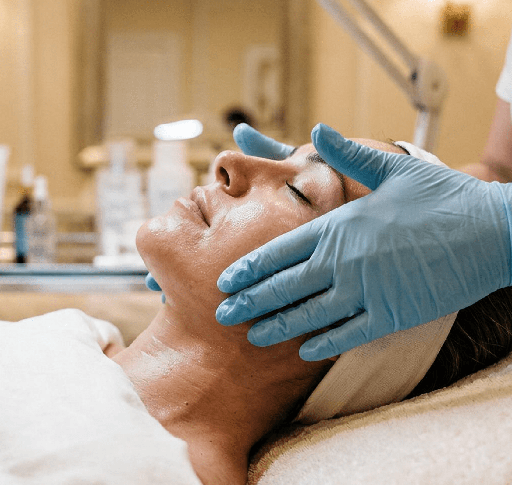 A clinician in blue gloves performing a facial treatment on a reclined patient