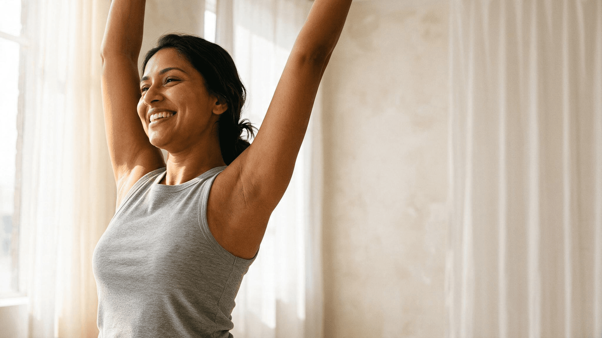 A smiling woman stretching both arms overhead in soft natural light