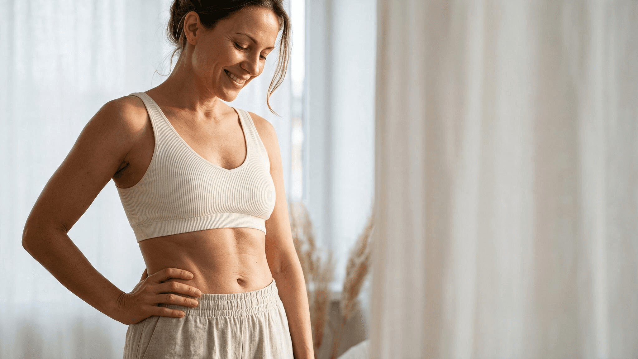 A smiling woman in a cream sports bra standing with a hand on her hip in soft daylight